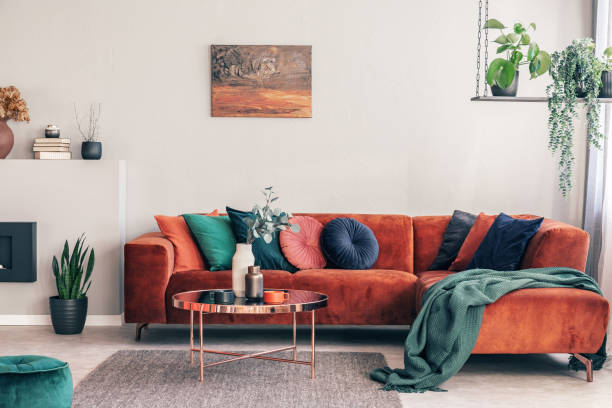 istock Stylish round table with flowers next to corner sofa with emerald pillows 1180794664
