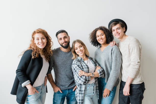 Smiling guys and girls in casualwear standing by white wall in front of camera Friendly smiling guys and girls in casualwear standing by white wall in front of camera and looking at you five people photos stock pictures, royalty-free photos & images