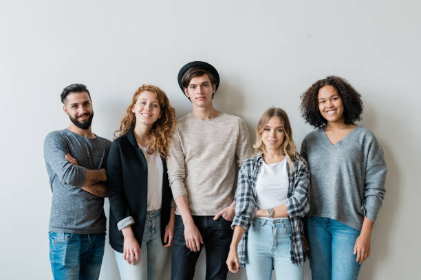 Cheerful intercultural friends in stylish casualwear looking at you Cheerful intercultural friends in stylish casualwear looking at you while standing against white wall in studio organized group photos stock pictures, royalty-free photos & images