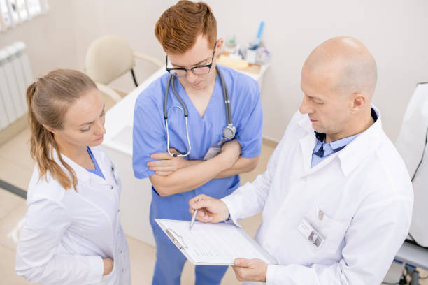 istock Bald serious doctor pointing at document while reading it aloud to young interns 1180815653