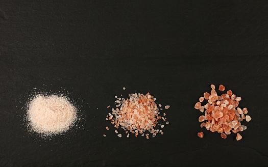 Close up three different grind size pink red Himalayan salt heaps over background of black slate board with copy space, elevated top view, directly above
