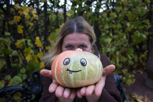 A blonde Caucasian woman and a pumpkin in her hands with a funny face drawn with a black felt tip pen in the Park on an autumn day