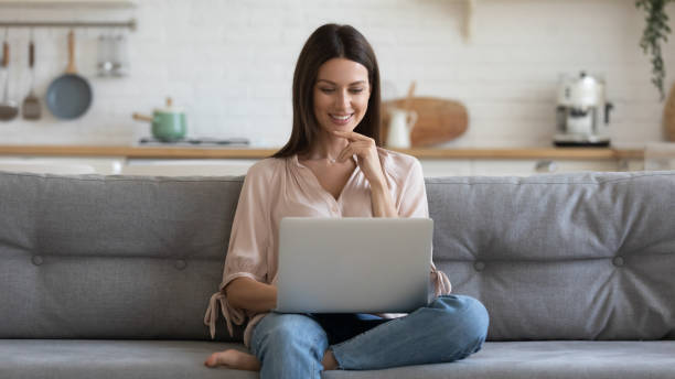 mulher nova de sorriso que usa o portátil, sentando-se no sofá em casa - web browsing - fotografias e filmes do acervo