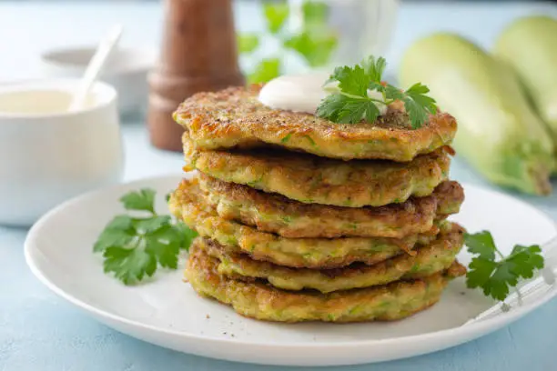 Zucchini fritters with fresh parsley and sour cream in plate on blue concrete background. Selective focus.