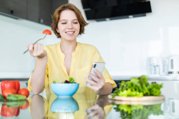 istock Pretty young woman using her mobile phone while eating salad in the kitchen at home. 1180841707