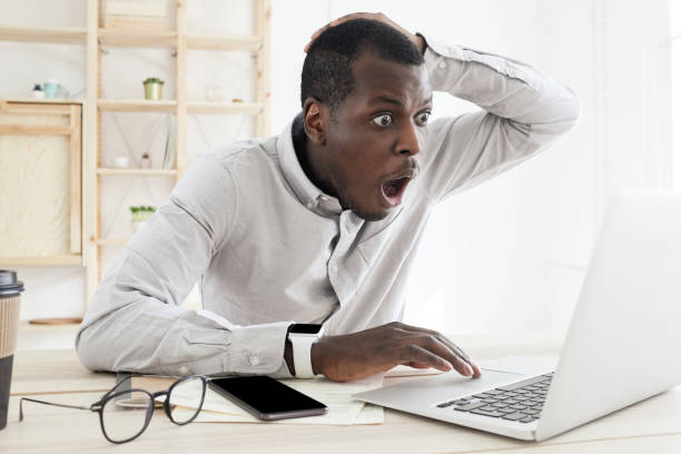 frustrado sorprendido hombre africano que tiene problemas, se siente confundido mirando la pantalla de la computadora portátil en la oficina - shock surprise people business fotografías e imágenes de stock