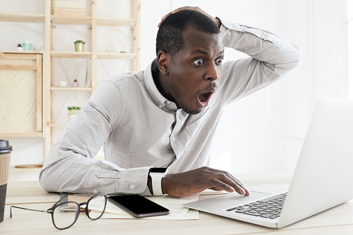 Frustrated shocked african man having problems, feel confused looking at laptop screen at office