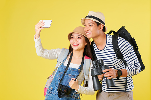Positive young Asian couple with backpack and binoculars talking selfie on smartphone when on vacation