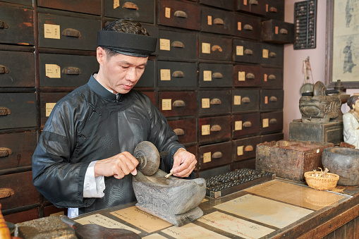 Serious mature traditional Asian medicine practitioner milling treatment ingredients in wooden boat tool