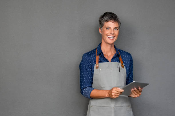 Mature woman in apron holding digital tablet Successful mature woman in apron standing and holding digital tablet against grey wall. Happy small business owner holding tablet and looking at camera. Smiling portrait of entrepreneur standing satisfied with copy space. cafeteria worker photos stock pictures, royalty-free photos & images