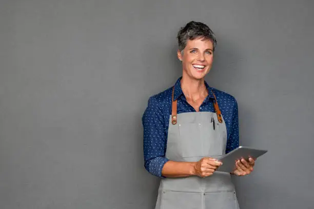 Successful mature woman in apron standing and holding digital tablet against grey wall. Happy small business owner holding tablet and looking at camera. Smiling portrait of entrepreneur standing satisfied with copy space.