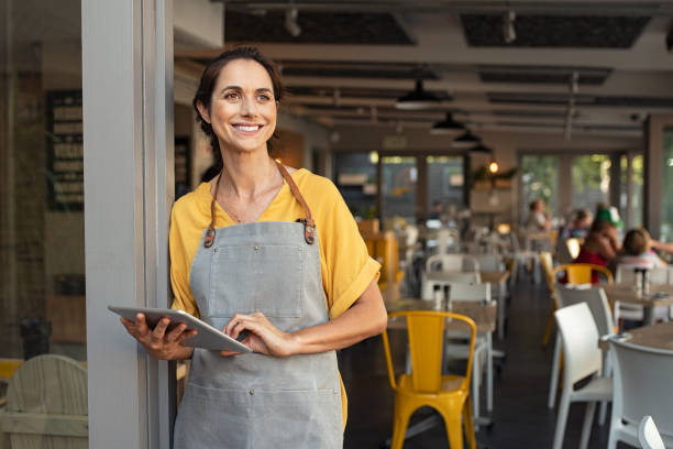 Successful owner standing at cafe entrance Portrait of a happy waitress standing at restaurant entrance holding digital tablet. Portrait of mature business woman standing at cafe entrance. Happy mature woman owner in grey apron standing at coffee shop entrance leaning while looking away with copy space. cafeteria photos stock pictures, royalty-free photos & images