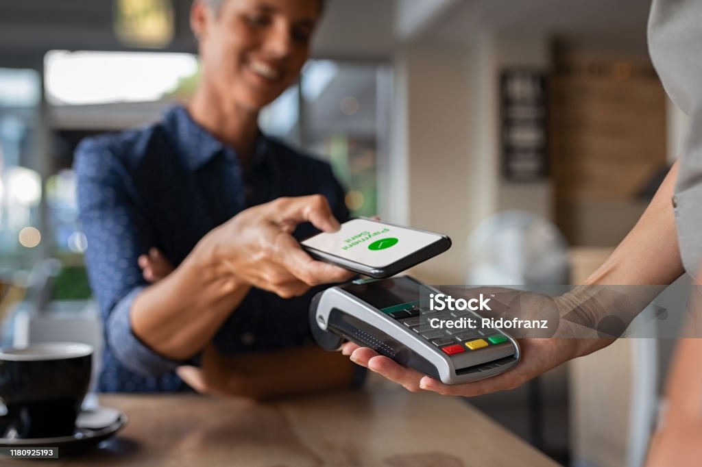 Woman paying using NFC technology Mature woman paying bill through smartphone using NFC technology in a restaurant. Satisfied customer paying through mobile phone using contactless technology. Close up hands of mobile payment at a coffee shop. Paying Stock Photo