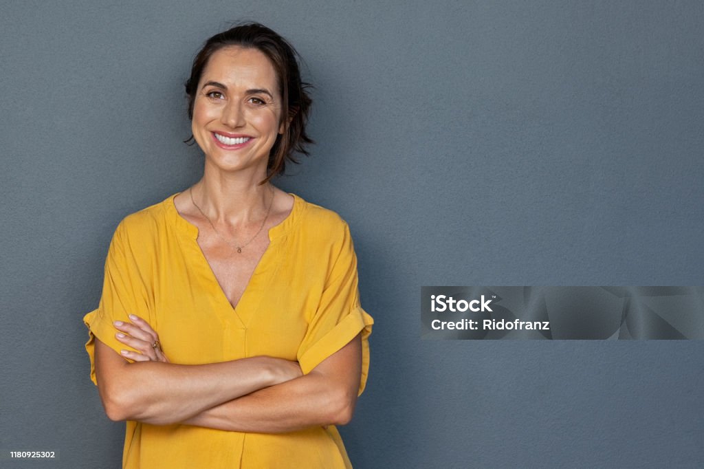 Happy mature woman smiling on grey wall Confident mature woman with crossed arms in casual clothing standing against grey background with copy space. Successful smiling woman with toothy smile looking at camera. Beautiful positive businesswoman standing. Women Stock Photo