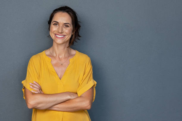 Happy mature woman smiling on grey wall Confident mature woman with crossed arms in casual clothing standing against grey background with copy space. Successful smiling woman with toothy smile looking at camera. Beautiful positive businesswoman standing. toothy smile photos stock pictures, royalty-free photos & images