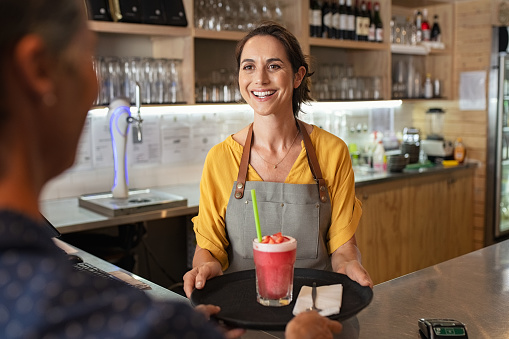 Happy waitress serving strawberry shake to customer at counter in cafeteria. Beautiful mature woman in grey apron holding tray with fresh strawberry smoothie and pieces for garnish.  Portrait of cheerful cafeteria worker serving customer at coffee shop.