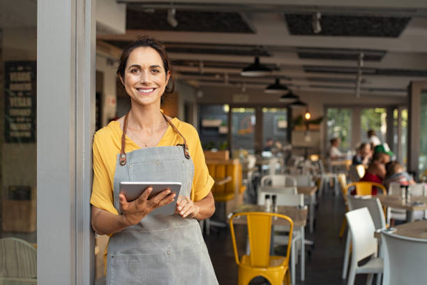 Small business owner at entrance looking at camera Portrait of happy woman standing at doorway of her store holding digital tablet. Cheerful mature waitress waiting for clients at coffee shop. Successful small business owner in casual clothing and grey apron standing at entrance and looking at camera. service occupation photos stock pictures, royalty-free photos & images