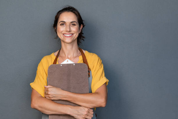 Woman entrepreneur in apron looking at camera Portrait of smiling mature woman in apron holding clipboard in hand while looking at camera. Beautiful waitress standing isolated against grey wall with copy space. Successful small business owner standing on grey background with copy space. cafeteria worker photos stock pictures, royalty-free photos & images