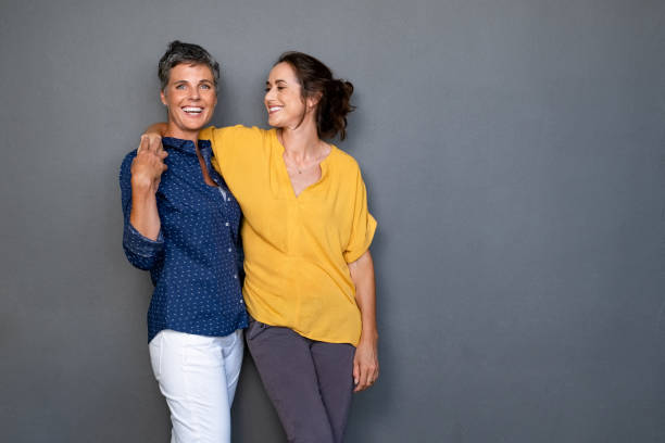 Mature friends women laughing together Mature happy women embracing each other against grey wall with copy space. Happy laughing ladies in smart casual standing on gray background. Cheerful middle aged woman with hand on shoulder of her stylish friend. lesbian stock pictures, royalty-free photos & images