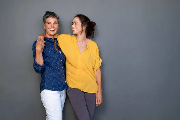 Mature happy women embracing each other against grey wall with copy space. Happy laughing ladies in smart casual standing on gray background. Cheerful middle aged woman with hand on shoulder of her stylish friend.