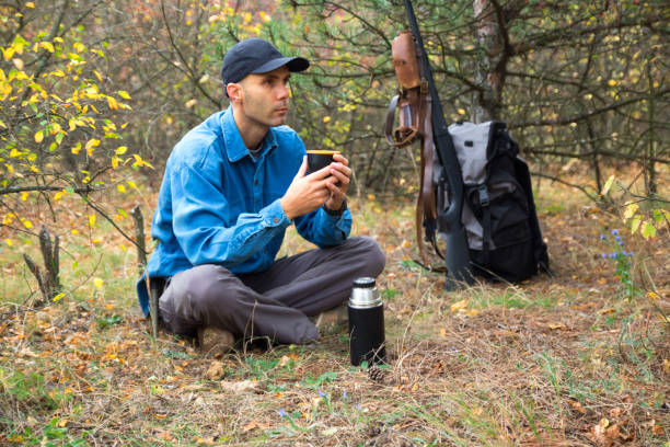 istock caucasian male hunter resting in forest. drinking tea from thermos. shotgun, ammunition, backpack on the background 1180930602