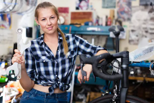 Young positive female is posing with instruments near bicycle in workshop.
