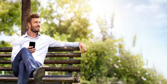 Portrait of young handsome business man in suit relax on bench in park. He holding smart phone and  watches thoughtfully in green forest park. Escape in nature to recover mind business concept.