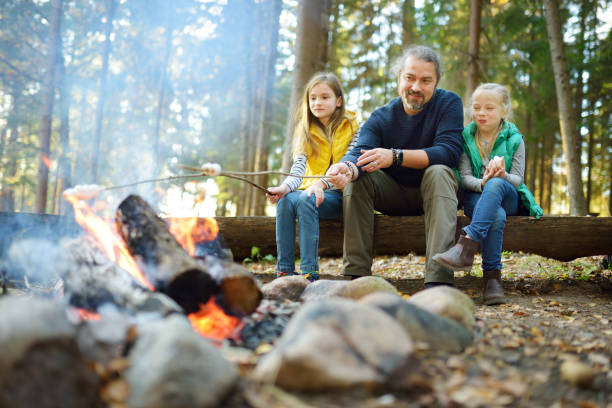 lindas hermanitas y su padre asando malvaviscos en palos en la hoguera. niños divirtiéndose en el campamento. camping con niños en el bosque de otoño. - camping family vacations eating fotografías e imágenes de stock