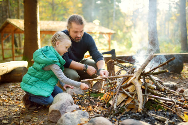 cute young girl learning to start a bonfire. father teaching her daughter to make a fire. child having fun at camp fire. camping with kids in fall forest. - vacations two generation family caucasian friendship imagens e fotografias de stock