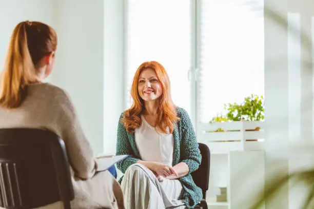 Mature woman smiling while discussing with therapist. Psychotherapist is with female at community center. They are sitting on chairs.