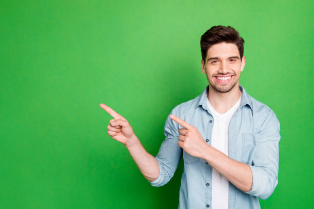 Photo of amazing salesman guy in excited mood indicating finger to empty space advising cool shopping prices wear casual denim shirt isolated green color background Photo of amazing salesman guy in excited mood indicating finger to empty space, advising cool shopping prices wear casual denim shirt isolated green color background one young man only stock pictures, royalty-free photos & images