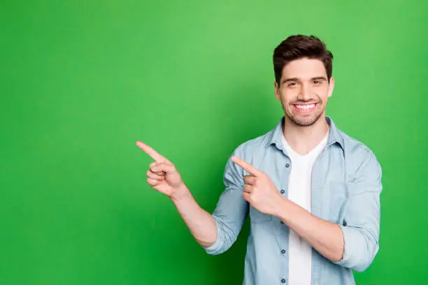 Photo of amazing salesman guy in excited mood indicating finger to empty space, advising cool shopping prices wear casual denim shirt isolated green color background