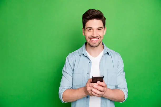 Photo of cheerful brown haired handsome blogger browsing through his telephone, looking for ideas to post in his social media isolated over green vivid color background