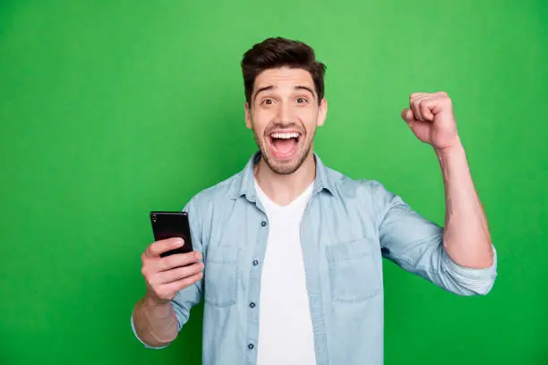 Photo of casual cheerful brunette haired crazy ecstatic overjoyed man rejoicing when having known of being accepted to job holding, phone shouting victoriously isolated over green vivid color background