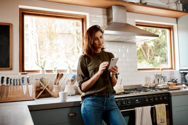 Let me respond to a few texts Cropped shot of an attractive young woman using a smartphone while standing in her kitchen at home house phone stock pictures, royalty-free photos & images