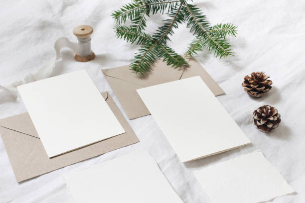 istock Christmas table, festive styled composition. Christmas greeting cards, envelopes mock-ups. Silk ribbon, pine cones and fir tree branch on white linen background. Top view. Festive craft, stationery. 1180954142