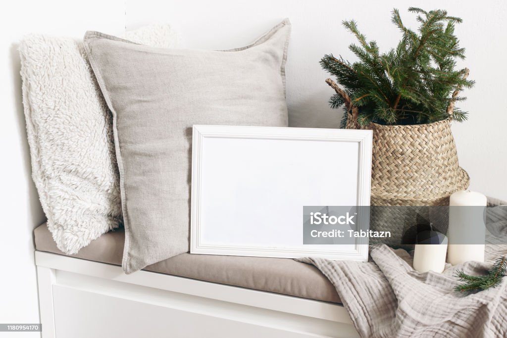 White blank wooden frame mockup with Christmas tree, candles, linen cushions and plaid on the white bench. Poster product design. Scandinavian home decor, nordic design. Winter festive concept. White blank wooden frame mockup with Christmas tree, candles, linen cushions and plaid on the white bench. Poster product design. Scandinavian home decor, nordic design, winter festive concept. Picture Frame Stock Photo