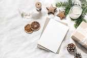 Christmas blank greeting card mock-up scene. Festive winter wedding composition. Craft envelope, pine cone, gift box, orange fruit slices and fir tree branch on white table, linen background. Flat lay.