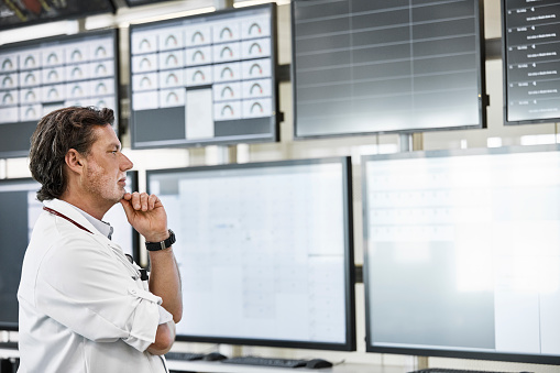 Thoughtful male medical professional doctor looking at device screens. Serious mature man is with hand on chin at medical clinic. He is wearing lab coat.