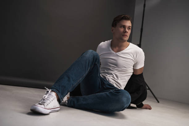 istock Young stylish guy posing in the studio on a gray background. Dressed in a white shirt, blue jeans and white sneakers. Model test 1180956809