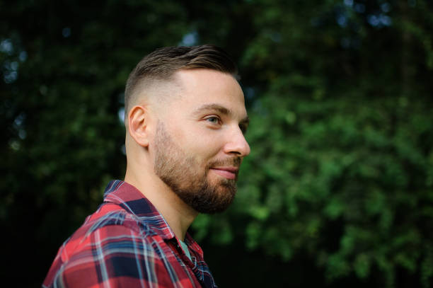 istock Brutal bearded young man in red shirt is standing outdoors on green trees background. Hipster style concept. 1180961349