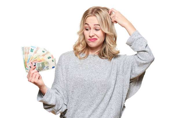 Pretty young woman in grey sweater holding bunch of Euro banknotes, scratching her head thinking, isolated on white background. stock photo