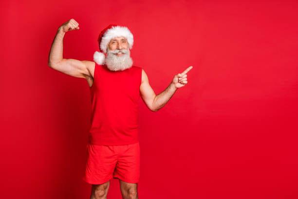 빨간 배경 위에 고립 된 스포츠 착��용을 입고 광고를 가리키는 이두근을 보여주는 흰색 머리 수염모자를 쓴 강력한 코치 산타 클로스의 초상화 - men gray hair vitality healthy lifestyle 뉴스 사진 이미지