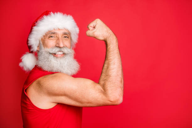 흰색 헤어 스타일과 모자 모자에 쾌활한 산타 클로스의 사진을 닫습니다, 빨간색 배경 위에 고립 된 스포츠웨어를 입고 자신의 팔뚝 체육관을 보여주는 - men gray hair vitality healthy lifestyle 뉴스 사진 이미지