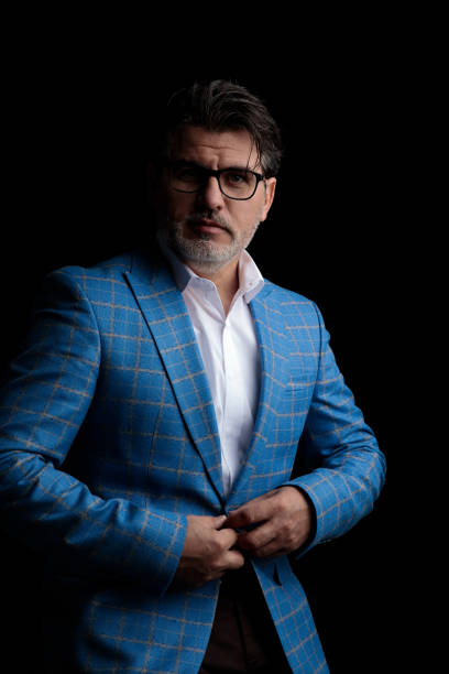 sexy formal business man wearing blue suit and glasses standing and holding hands on jacket's button while staring at camera serious against black studio background