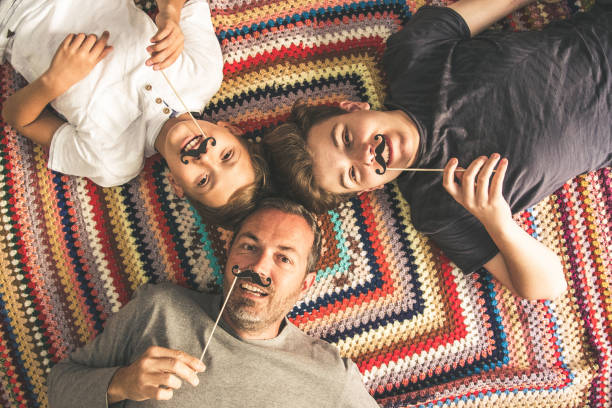 Father and two sons enjoying together lying on a colorful blanket. Tree men of different ages smiling playing with fake mustache. Top view of a couple of teen and their dad. Father and two sons enjoying together lying on a colorful blanket. Tree men of different ages smiling playing with fake mustache. Top view of a couple of teen and their dad. happy fathers day funny stock pictures, royalty-free photos & images