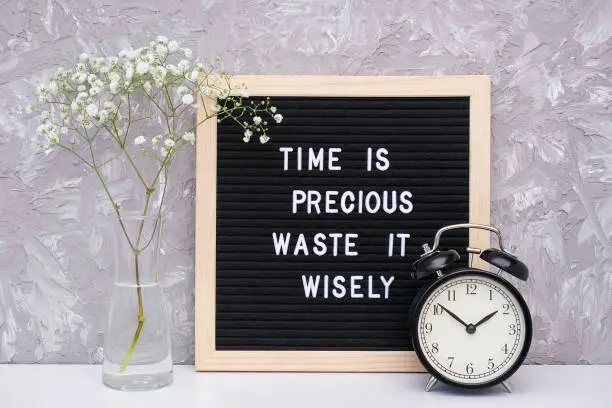 Time is precious waste it wisely. Motivational quote on letterboard, black alarm clock, flower in vase on table against stone wall. Concept quote of the day.