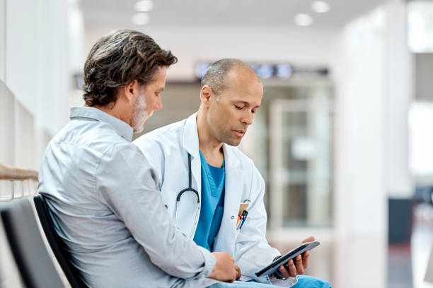 Male Doctor Counseling Mature Patient At Hospital Male doctor using digital tablet while counseling mature patient in waiting room. Mature is discussing with medical professional in hospital. They are sitting on seat. scandinavian ethnicity stock pictures, royalty-free photos & images