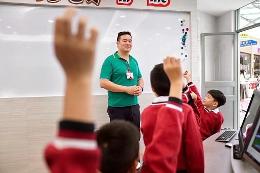 Male teacher looking at elementary students in computer lab. School children are raising their hands in classroom. Professional is teaching pupils.