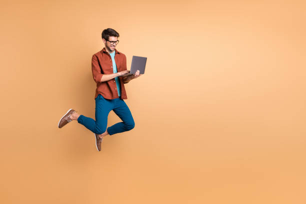 full length body size view of his he nice attractive cheerful cheery successful brunet guy jumping in air using laptop home-based job isolated over beige color pastel background - job joy student computer imagens e fotografias de stock